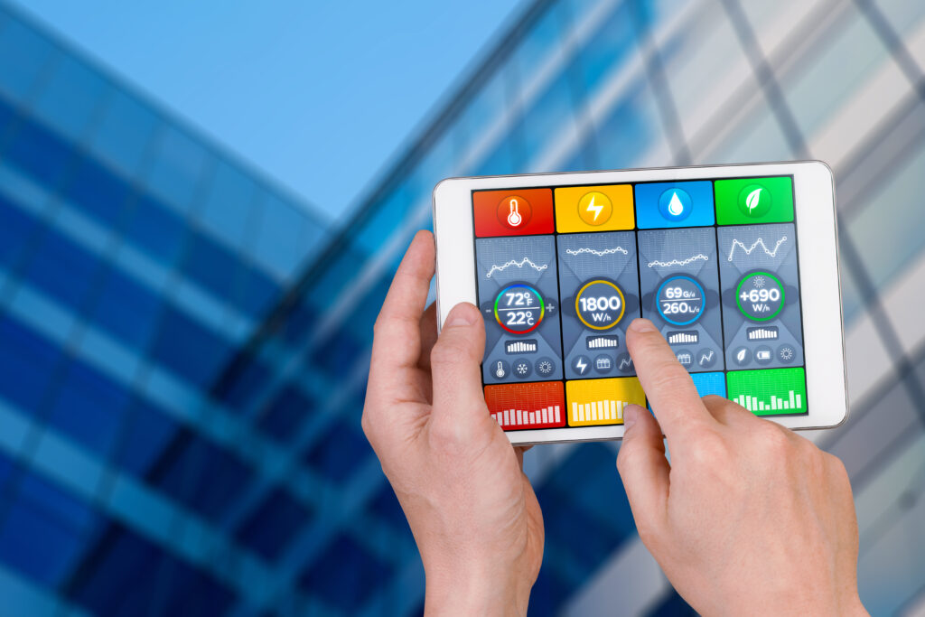 Intelligent building automation: hand pointing an interactive tablet display showing building consumptions related to temperature and heating, power and light, water usage and green energy production. With charts, data and interactive functions for remote control of building facilities and utilities.
